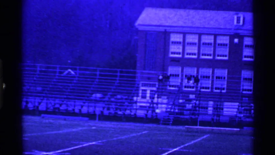 1969 Ohio 8mm frame showing a blue-tinted school sports field with bleachers and a brick building behind it.