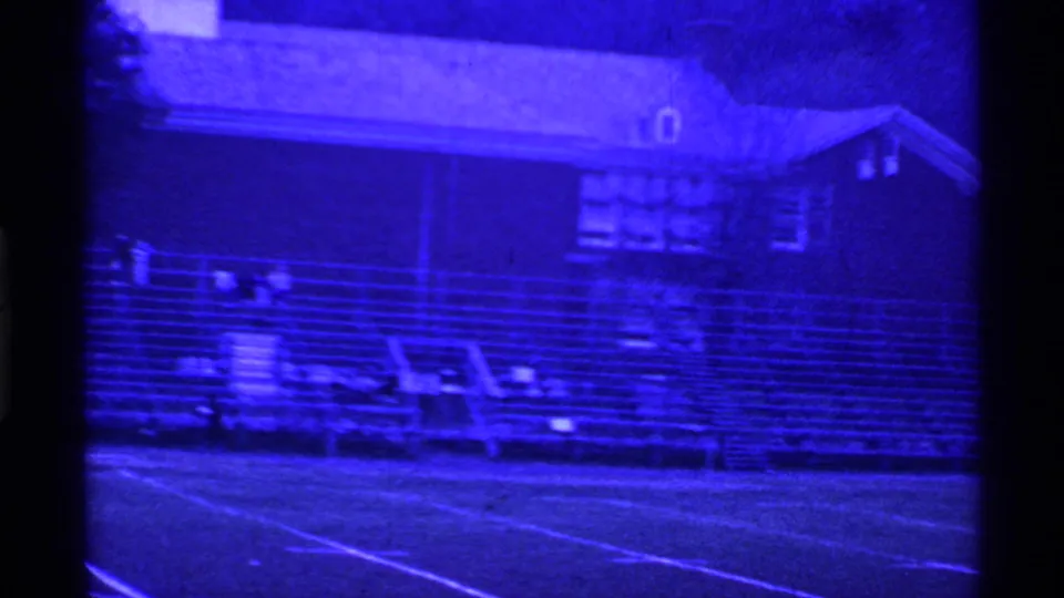 1969 Ohio 8mm frame showing bleachers, field markings, and a brick school building under a strong blue cast.