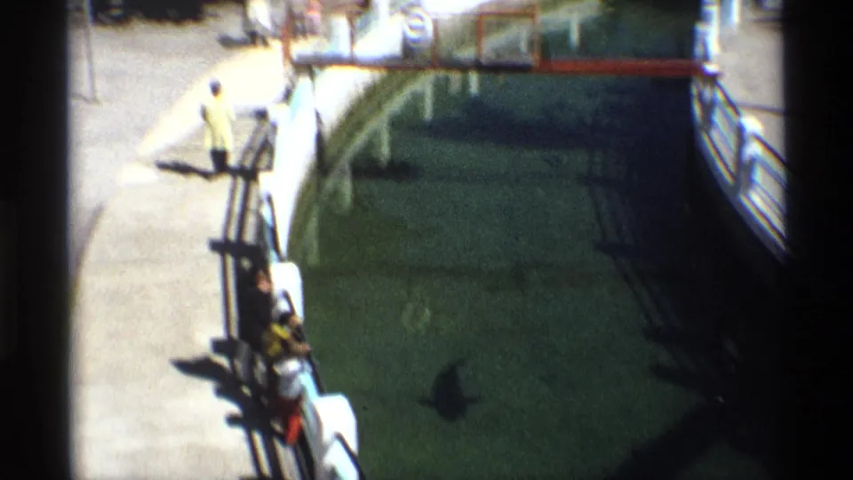 Visitor in a yellow shirt stands near the railing at a large aquarium tank in archival 8mm footage.