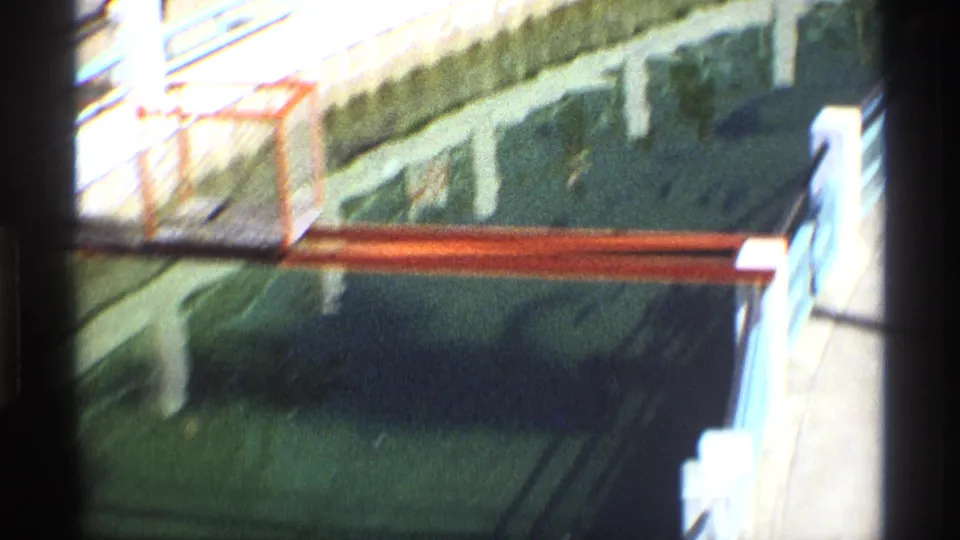 1970 8mm frame showing dark water under a bridge, with a red railing, white concrete posts, and heavy shadows along the film edge.