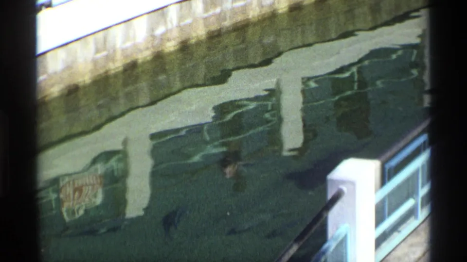 1970 8mm frame with a large dark fish-like or whale-like form just below the surface near reflected bridge supports and a white rail post.