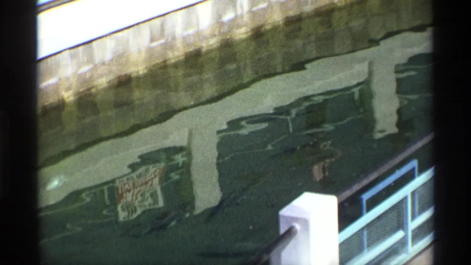 1970 8mm view of rippling water beneath a concrete bridge, with railings at right and a dark aquatic form partly visible near the bottom.