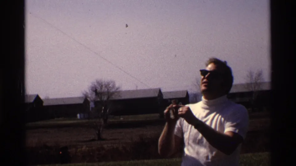 1970 home-movie frame from Florida or Michigan showing an adult man in sunglasses handling a kite line in a rural field under blue sky.