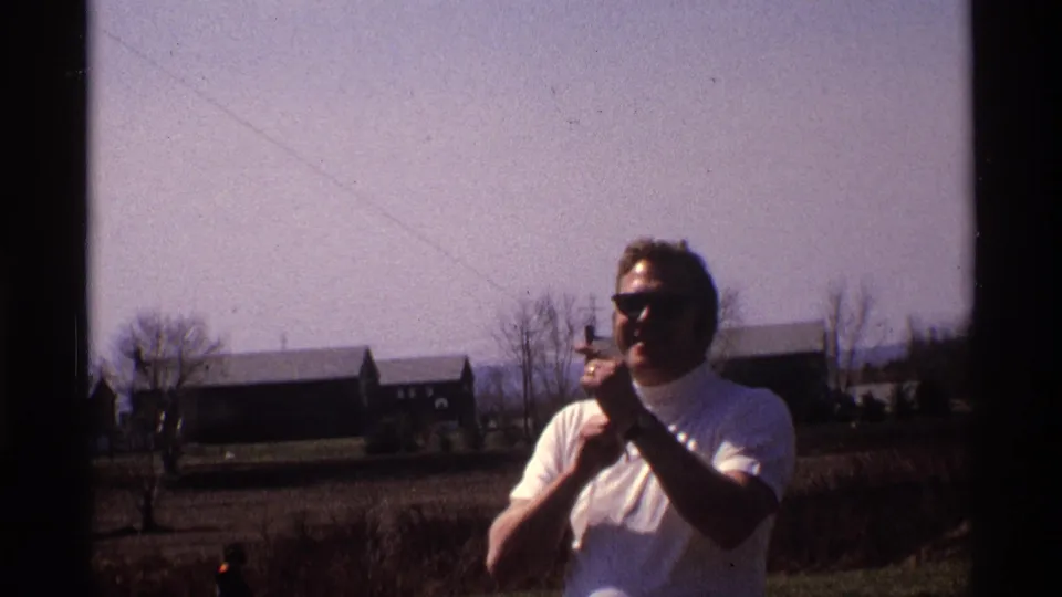 The 1970 Florida, Michigan home-movie frame shows the man holding kite string, with bare trees and low buildings across the field.