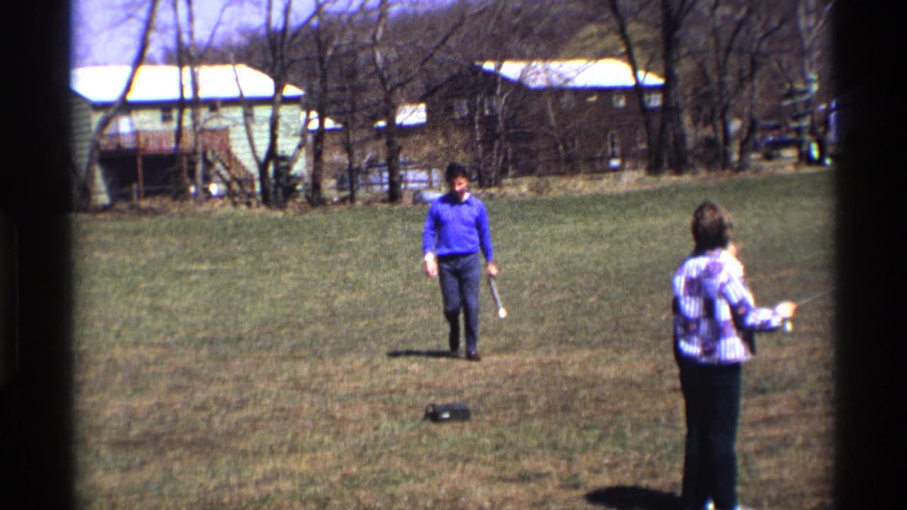 Man in a blue sweater walks across a grassy yard toward a person holding kite string.