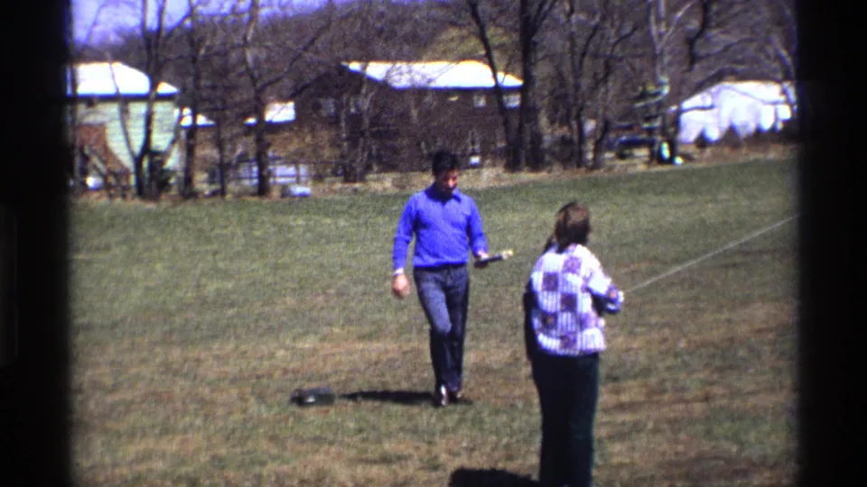 In the 1970 home-movie frame, the man nears the kite flyer while a pale kite line cuts across the grassy residential setting.