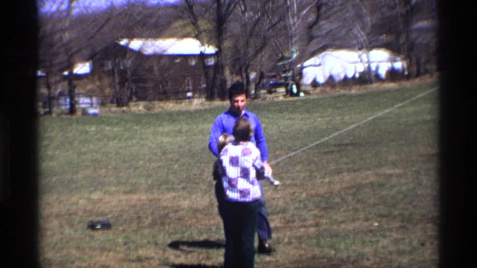 The man reaches the kite flyer in this metadata-supported 1970 Florida, Michigan yard scene, with houses and leafless trees in the background.