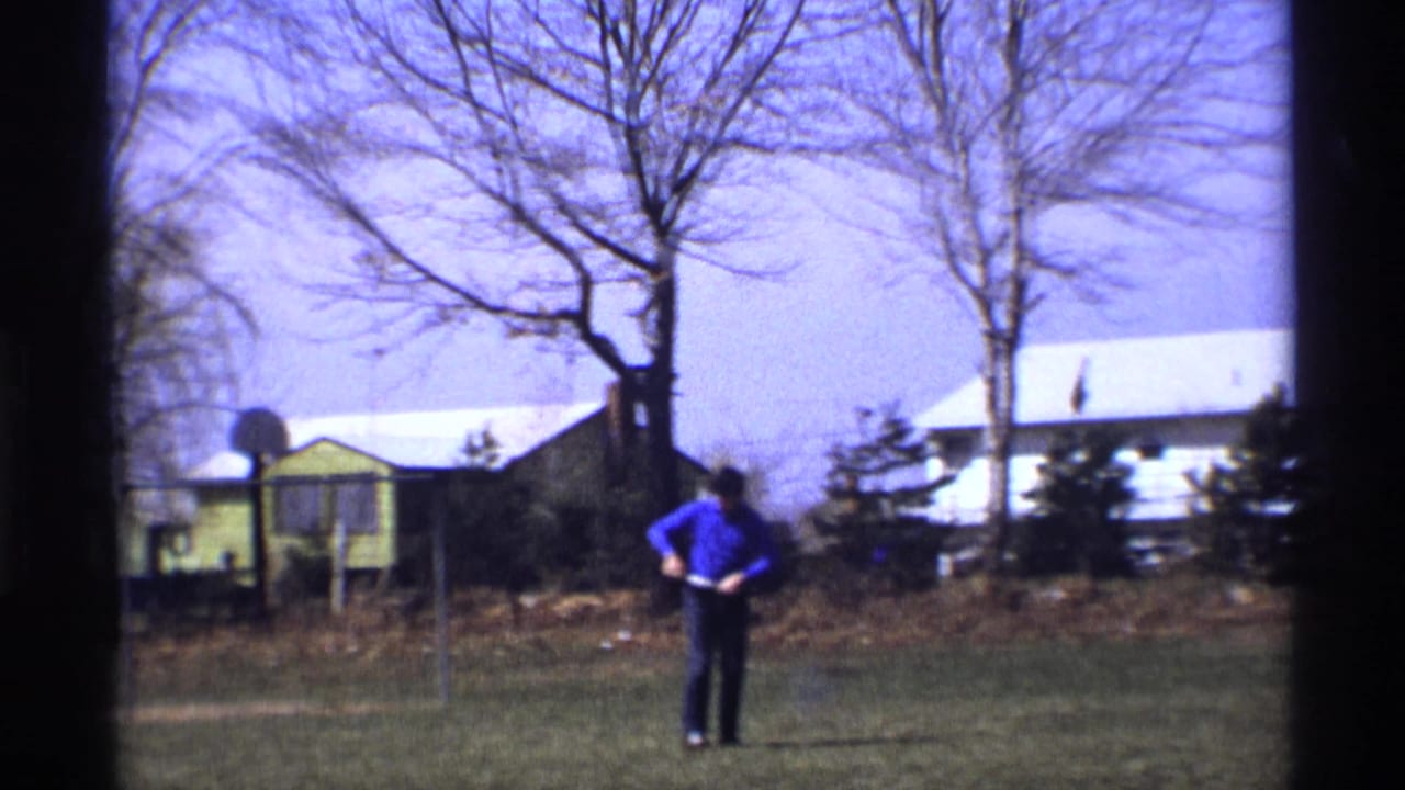 Man in a blue shirt stands in a grassy yard with bare trees and houses behind him.