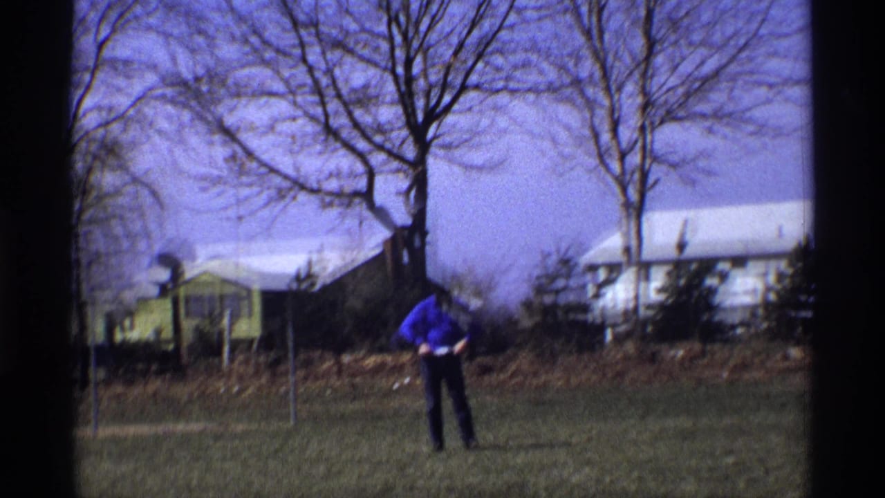 Man stands farther back on a lawn with a yellow house, bare trees, and shrubs behind him.