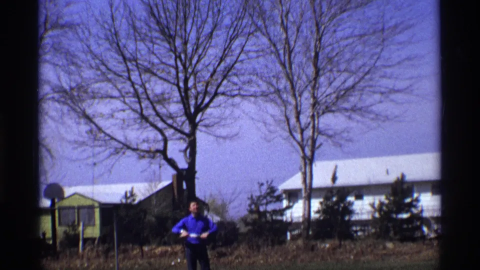 The 1970 home-movie frame shows a man low in the shot, standing in a yard below bare trees and houses.