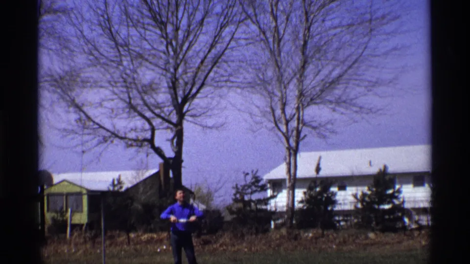 A man in a blue shirt holds a light object in a residential Florida, Michigan yard, with bare trees and houses behind him.