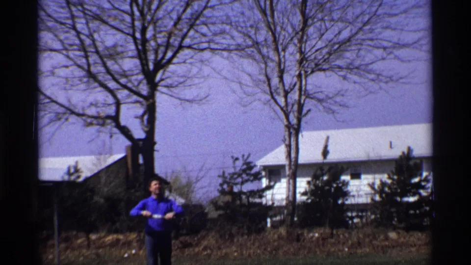 In supported 1970 Florida, Michigan footage, a man in a blue shirt stands in a suburban yard holding kite string.
