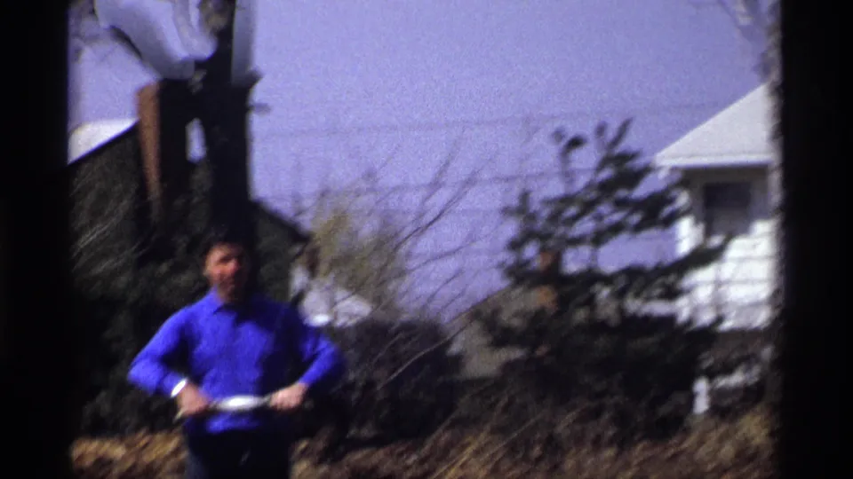 The man stands closer to camera in the suburban yard, holding a kite string winder against a pale sky.