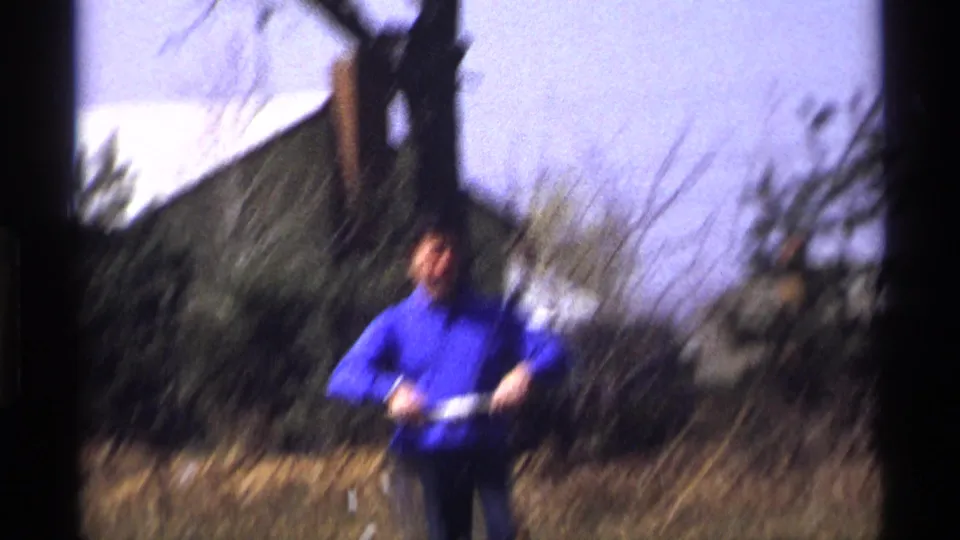 A soft-focused 8mm frame shows the man in blue holding the winder in the backyard, with houses and trees behind him.