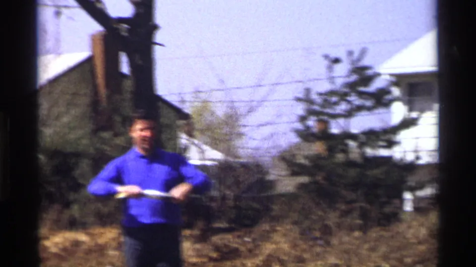 The man remains centered in the 1970 Florida, Michigan yard scene, holding the kite string winder near trees and houses.