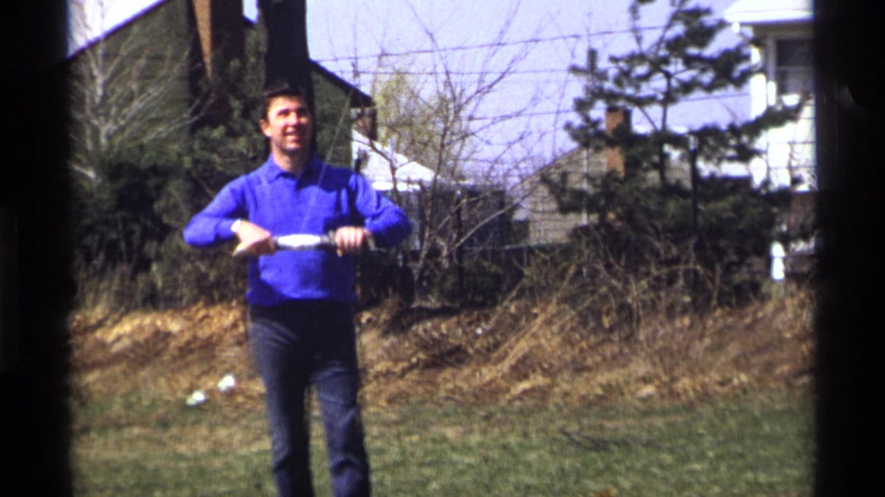 Man in a blue shirt stands in a yard holding a roll of string or tape.