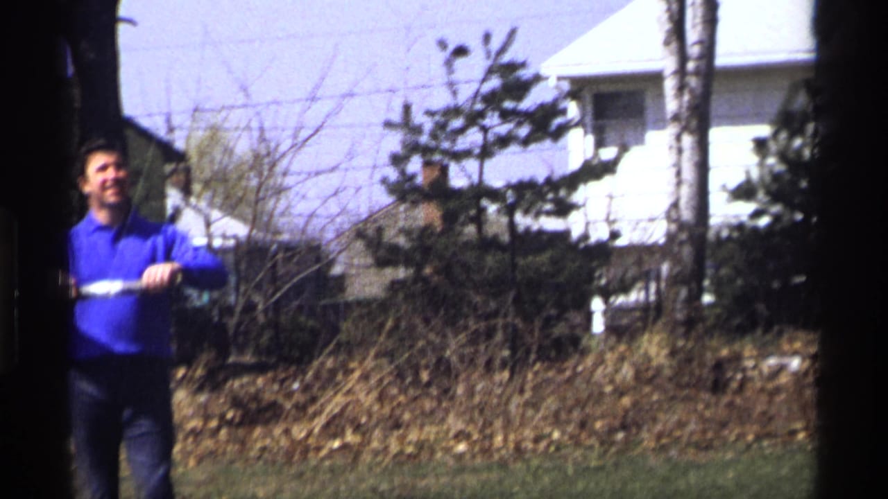 Man in a blue shirt stands near a chain-link fence with houses behind him.