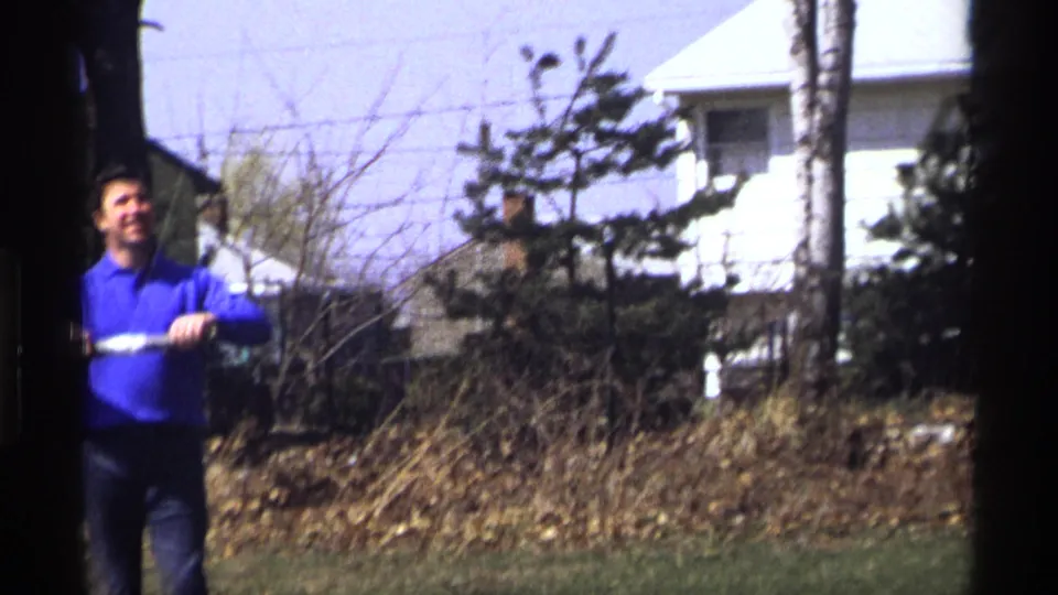 The man remains in the yard, partly framed by film gate edges, with a chain-link fence, shrubs, and houses visible behind him.