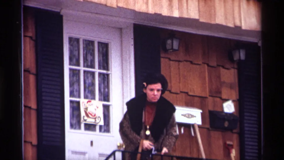 Adult woman stands on a 1970 New York City stoop, wearing a dark vest over patterned sleeves near a decorated front door.