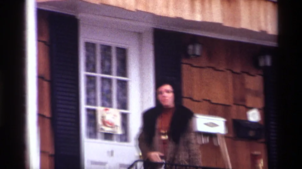 Blurred 8mm frame of the woman shifting on the New York City stoop, with the white door and holiday decoration still visible.