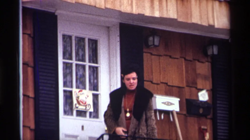 Adult woman on a 1970 New York City residential stoop, framed by black shutters, wood shingles, and a Santa door decoration.