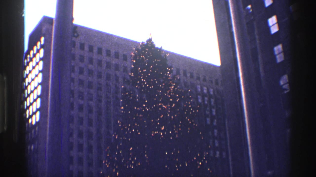 Lit Christmas tree framed by tall buildings and pale sky.