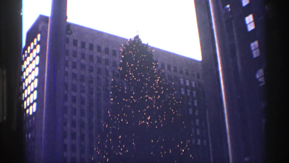 1970 8mm view of the Rockefeller Center Christmas tree glowing between dark New York City office buildings.