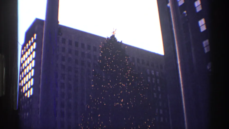 Visitor-shot 8mm frame of the Rockefeller Center Christmas tree with a star topper between high-rise facades.