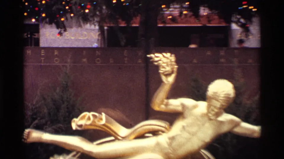 1970 Rockefeller Center plaza frame showing a golden statue in the foreground with holiday lights and tree decorations behind it.