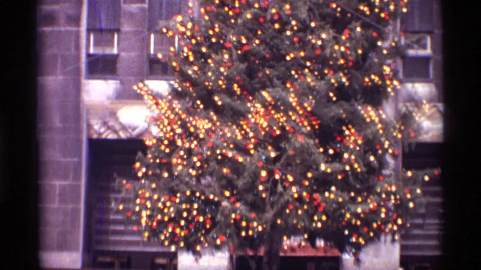 A large illuminated Rockefeller Center Christmas tree dominates the frame against a New York City stone skyscraper facade.