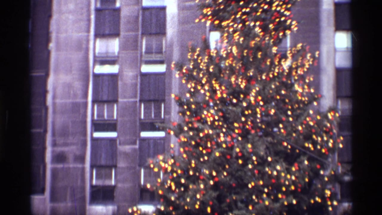 1970 New York holiday home-movie frame showing a lit Christmas tree against a vertical stone skyscraper facade.
