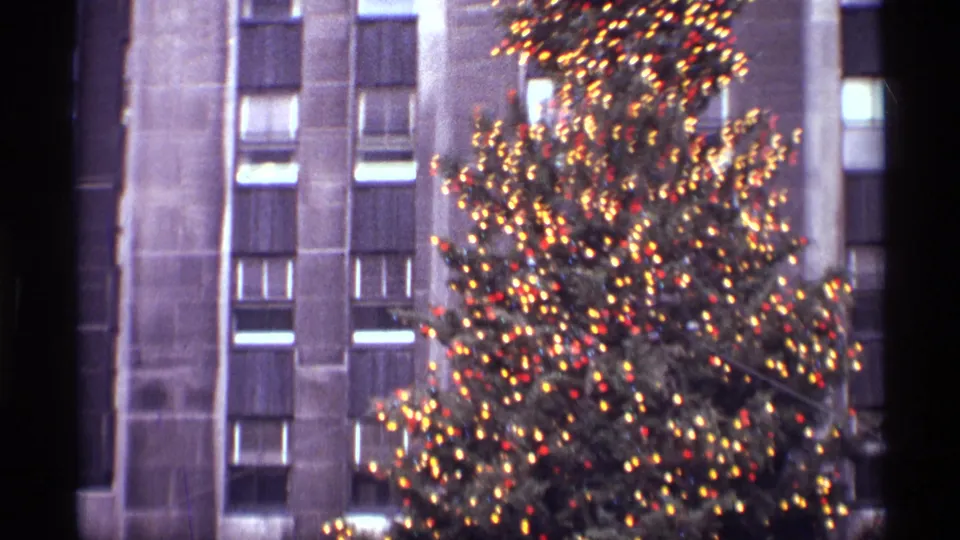 1970 New York holiday home-movie frame showing a lit Christmas tree against a vertical stone skyscraper facade.