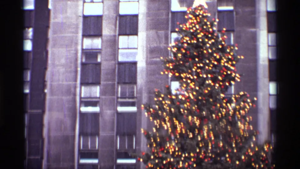 1970 New York frame with the illuminated Rockefeller Center Christmas tree offset beside a stone high-rise facade.