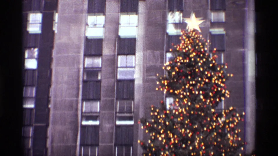 1970 New York holiday frame showing the star-topped Rockefeller Center Christmas tree before tall Art Deco-style windows.