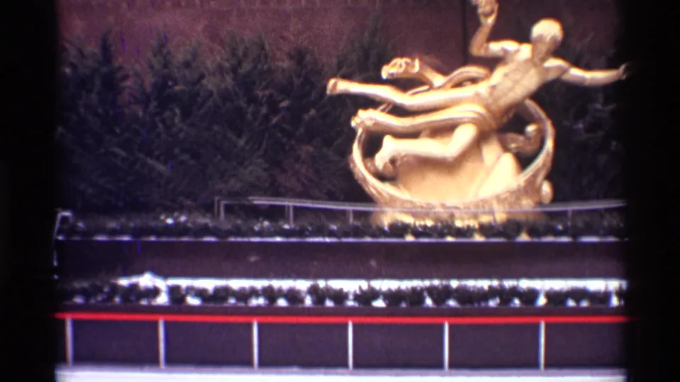 1970 New York City 8mm frame with a gold outdoor sculpture, evergreen backdrop, railing, and modern building facade elements.