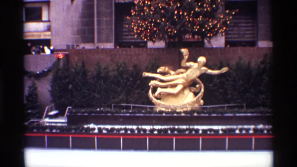 1970 New York City home-movie frame showing a golden sculpture, illuminated Christmas tree, evergreen shrubs, and a building entrance area.