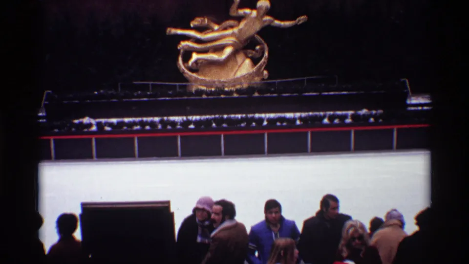 1970 8mm Rockefeller Center frame with bundled visitors near the rink edge and the gold Prometheus sculpture in the background.