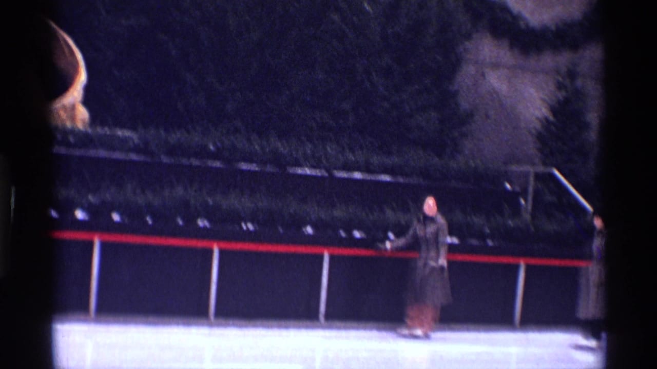 1970 New York City home-movie frame with a distant skater in winter clothing near the rail of an outdoor ice rink.