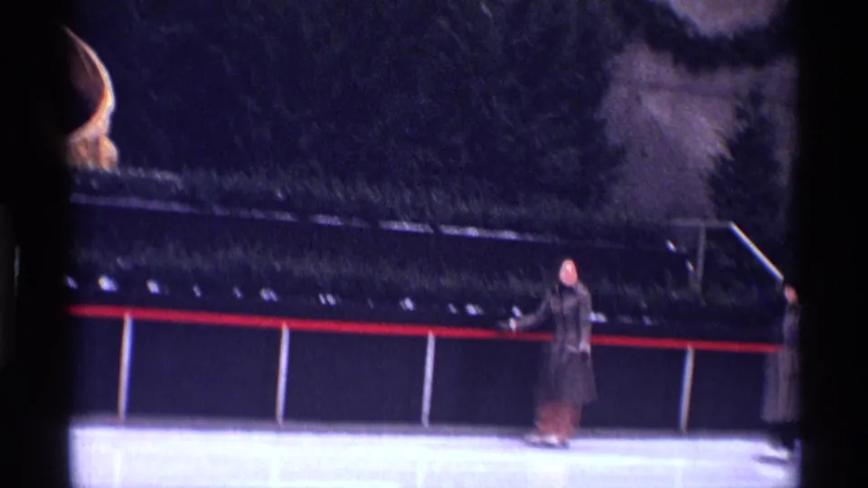 1970 New York City home-movie frame with a distant skater in winter clothing near the rail of an outdoor ice rink.