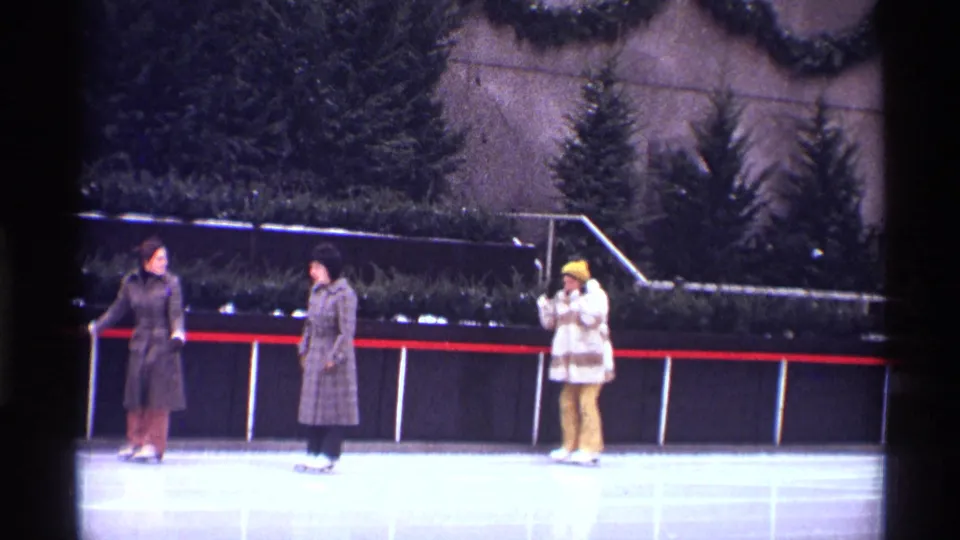 1970 New York City rink scene with two women near the rail and another person in a yellow hat holding a camera or binoculars.