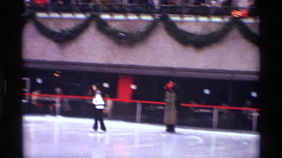 1970 New York City outdoor rink frame with two skaters, a red barrier, garland, and a lit Christmas tree in the background.