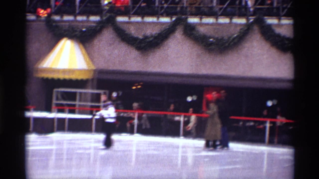 Skaters on the Rockefeller Center rink with garland, red railings, and a striped awning behind them.