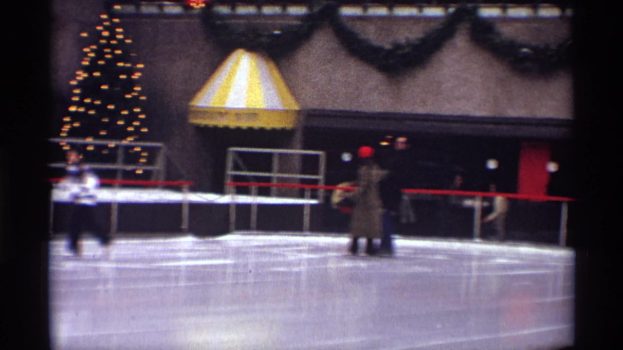 A couple skates near the center of the rink with a lit Christmas tree and striped awning in the background.