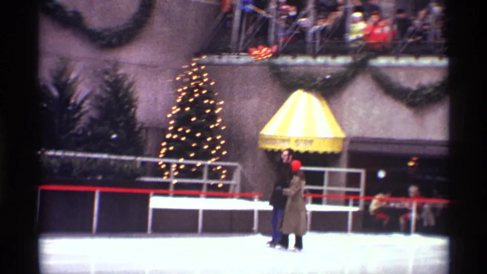 1970 8mm frame from Rockefeller Center showing two skaters near the rail, a lit Christmas tree, a striped awning, garland, and spectators watching from above.
