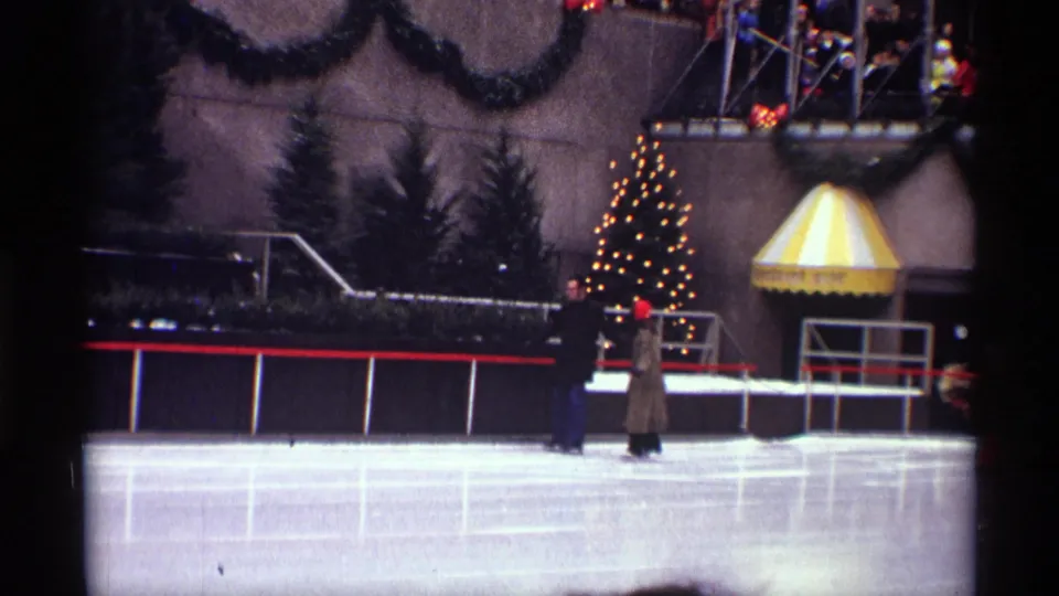Wide 1970 home-movie frame of the Rockefeller Center rink in New York City, with a skating couple, lit Christmas tree, decorated facade, spectators, red railings, and a striped awning.