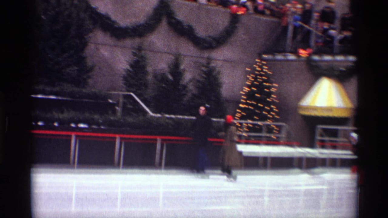 Outdoor ice rink with skaters, evergreen planters, a decorated Christmas tree, and spectators above the rink.
