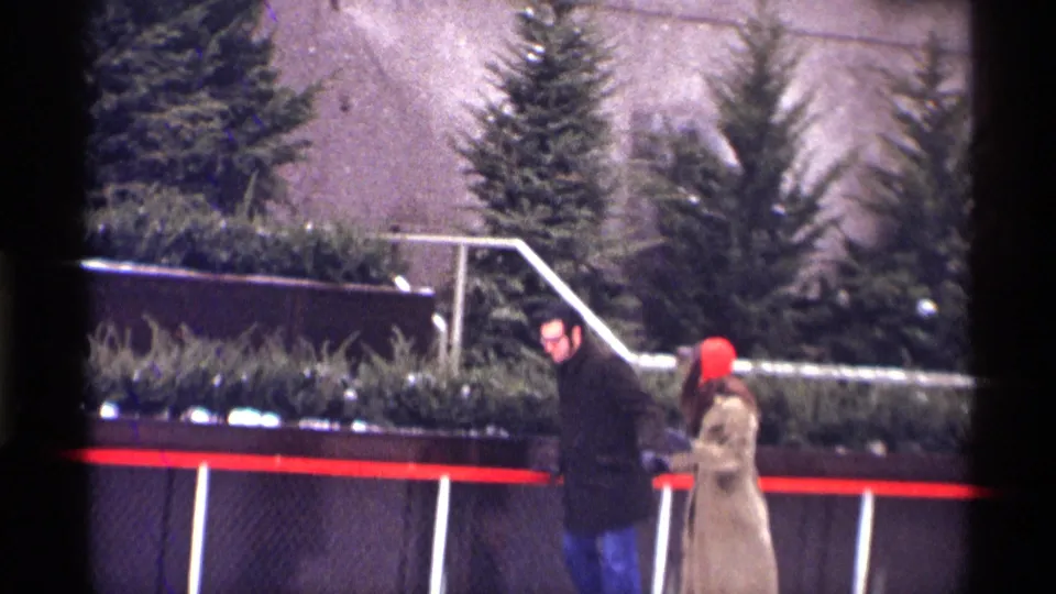 1970 New York City frame of a man in a dark coat and glasses beside a woman in a beige coat and red hat near an outdoor rink railing.