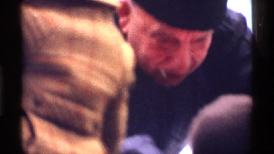 Close home-movie view of a bundled man in a dark winter hat among other visitors at the New York City skating scene.