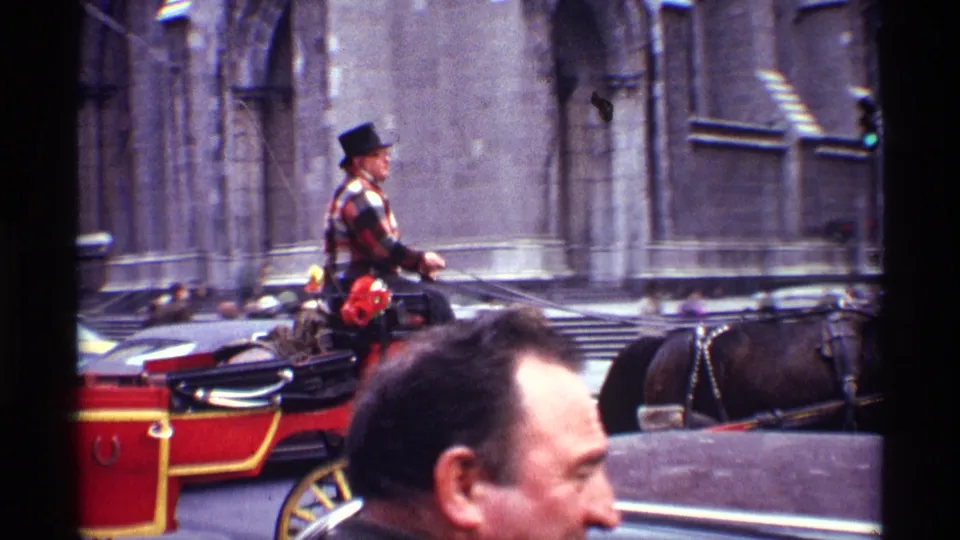 1970 New York City 8mm frame with the carriage driver, red carriage, horse harness, passing cars, and a pedestrian face in the foreground.