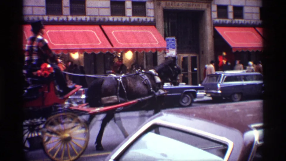A top-hatted carriage driver guides a dark horse along a 1970 New York City street beside classic cars and red awnings.
